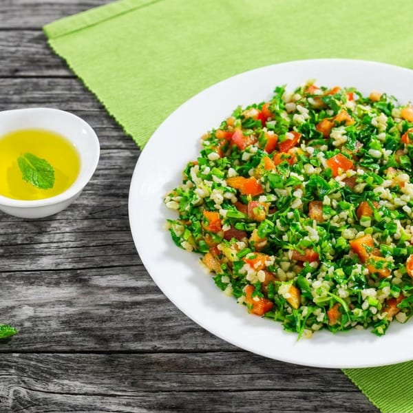 White plate of tabbouleh salad with bulgur, chopped greens, tomatoes, and carrots beside mint-infused oil on a wooden table.