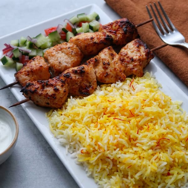 Grilled chicken skewers with saffron rice, cucumber salad, and yogurt sauce on a white platter.